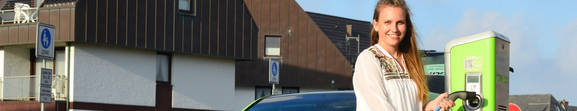 Frau steckt Ladekabel in Ladesäule, lächelt in die Kamera