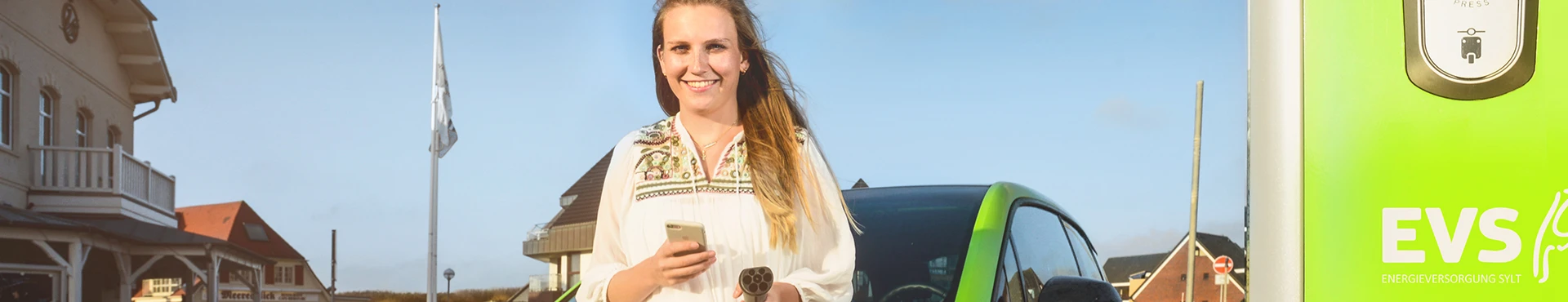Lächelnde Frau mit Smartphone steht mit E-Auto vor Ladesäule