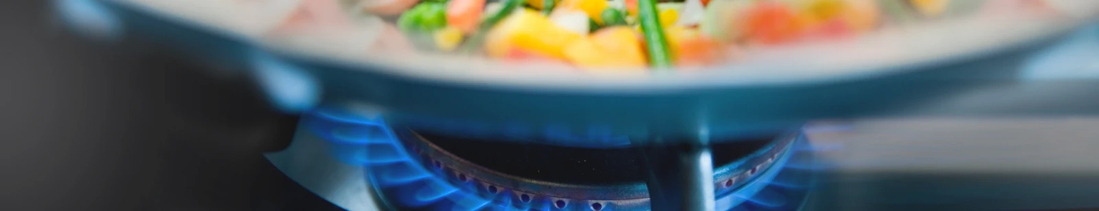 Nahaufnahme einer Gas-Herdflamme mit Topf in dem Gemüse erkennbar ist